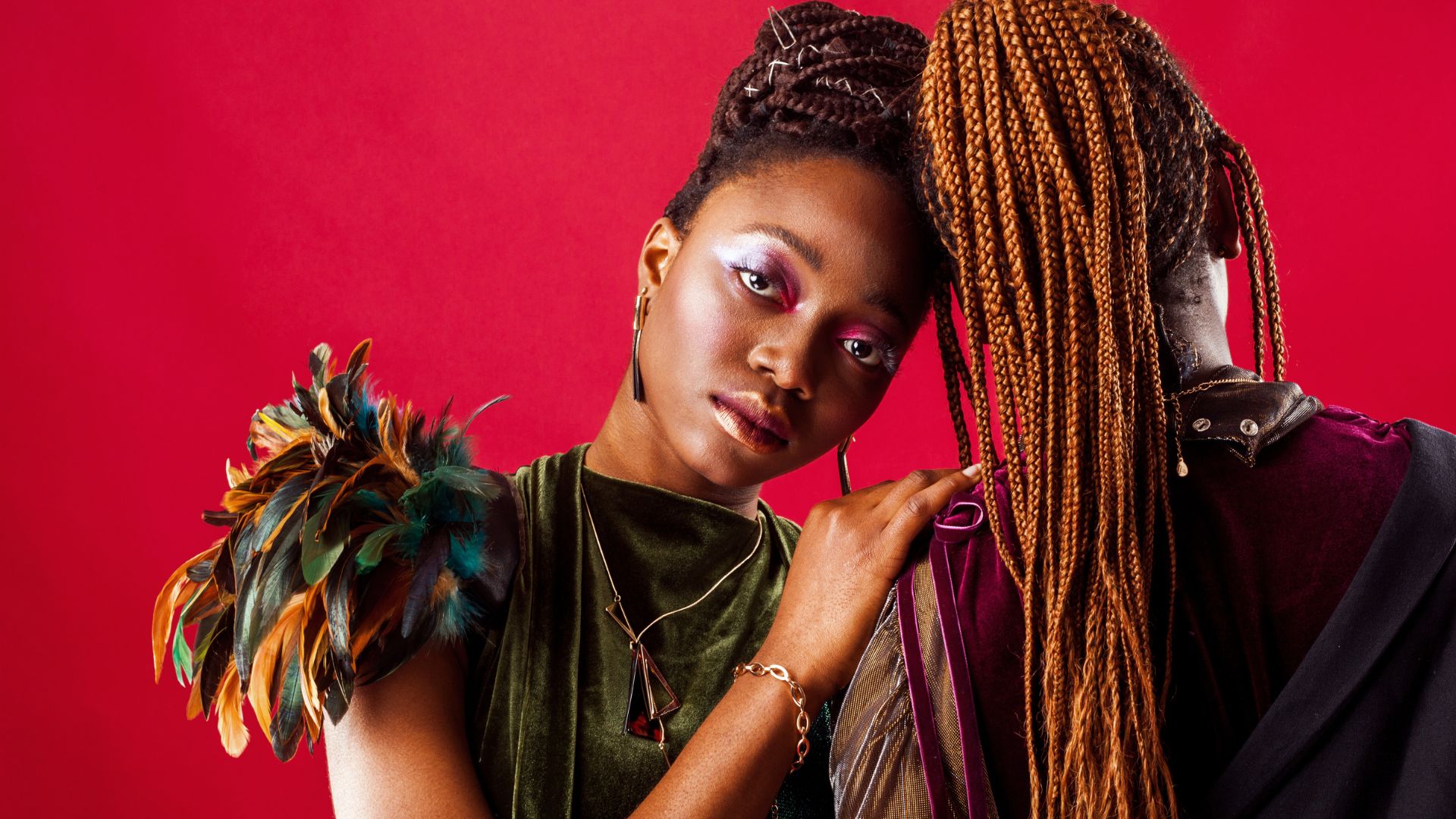 Woman with braids and feather accessory poses against vibrant red background